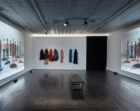 Installation image showing hanging jackets, bench, and wall projections of people standing